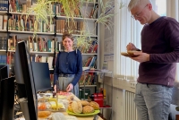 Blick in das Forschungsstellenbüro von ALMA. im Vordergrund ein Bildschirm auf einem Schreibtisch. Rechts vom Tisch steht ein Mann und isst ein Brötchen von einem Teller. Hinter dem Tisch steht eine Frau und lächelt in die Kamera. Hinter ihr ein Bücherregal. Von oben ragt eine Zimmerpflanze in das Foto hinein.