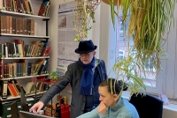 Blick in das Forschungsstellenbüro von ALMA. Im Vordergrund ein Bildschirm auf einem Schreibtisch. Dahinter sitzt eine Frau und schaut auf den Bildschirm. Links davon steht ein Mann mit Hut, Schal und Mantel bekleidet und zeigt auf den Bildschirm. Im Hintergrund ein Bücherregal. Rechts oben hängt eine Zimmerpflanze in einem braunen Blumenkübel von der Decke.