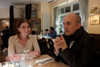 Ein Mann und eine Frau sitzen an einem Tisch im Restaurant. Die Frau sitzt links, der Mann an der Kopfseite des Tisches rechts von ihr. Vor ihnen auf dem Tisch ist eine Glaskaraffe mit Wasser zu sehen. Die Frau lächelt in die Kamera, der Mann blickt auf den Tisch vor sich. Im Hintergrund ist der Innenraum des im Landhausstil gehaltenen Restaurants zu sehen sowie einige Gäste an anderen Tischen. 