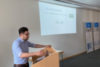 Gustavo Campedelli steht hinter einem Redepult. Seine Arme hat er darauf abgelegt. An der dahinterliegenden Wand wird eine PowerPoint angezeigt. Der Redner ist von der Seite her aufgenommen. 