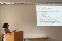 Ragini Menon steht hinter einem Redepult und hat die rechte Hand darauf abgelegt, die linke weist auf die PowerPoint-Präsentation, die an der dahinterliegenden Wand angezeigt wird.