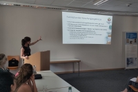Sabine Tittel steht hinter einem Redepult und weist mit einer Hand auf die PowerPoint-Präsentation, die an der dahinterliegenden Wand angezeigt wird. Im Vordergrund sind einige Mitglieder des Teams der Forschungsstelle "Bibelglossare" zu sehen.