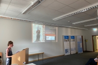 Viola Mariotti steht hinter einem Redepult und hat die Hände darauf abgelegt. An der dahinterliegenden Wand wird eine PowerPoint-Präsentation angezeigt, die ein Heiligenbild zeigt.