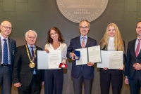 Sechs Personen stehen vor einer dunkelgrau gekachelten Wand. Die mittleren drei Personen, zwei Frauen und ein Mann, halten jeweils eine Urkunde und eine Medaille in der Hand.