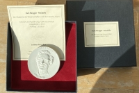 Eine geöffnete, graue Box auf hölzernem Untergrund. Ihr Inneres ist rot verkleidet und beinhaltet eine Medaille mit dem Konterfei eines Mannes, der eine Brille trägt. Auf der Medaille steht in Großbuchstaben „Kurt Ringger 1934 – 1988“ Es liegt ebenfalls ein Schein bei.