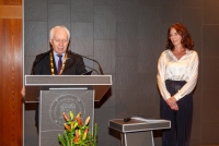 Eine Frau mit offenen, dunkelbraunen Haaren steht neben einem Podium. Sie trägt eine dunkle Hose und eine helle Bluse. Sie sieht zu einem Mann, der am Podium steht. Er trägt einen dunklen Anzug und hat eine goldene Kette um den Hals. Vor dem Podium steht ein orangenes Blumengesteck.