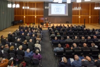 Ein mit Menschen gefüllter Plenarsaal. Auf der Bühne steht ein Mann am Podium und hält eine Rede.
