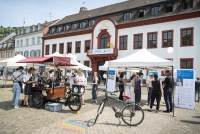 Im Vordergrund wird Kaffee ausgeschenkt und Menschen informieren sich an verschiedenen Ständen über Projekte des Wissenschaftlichen Nachwuchses. Im Hintergrund ist das Akademiegebäude.  
